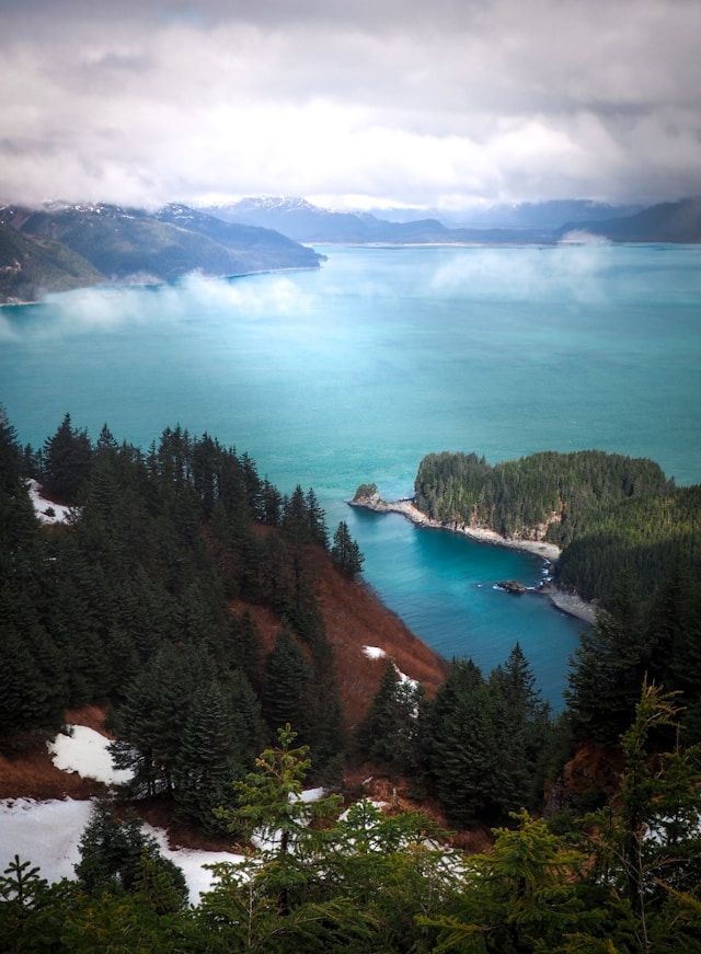 An elevated view of a turquoise bay surrounded by pine-covered mountains and patches of snow under a cloudy sky.