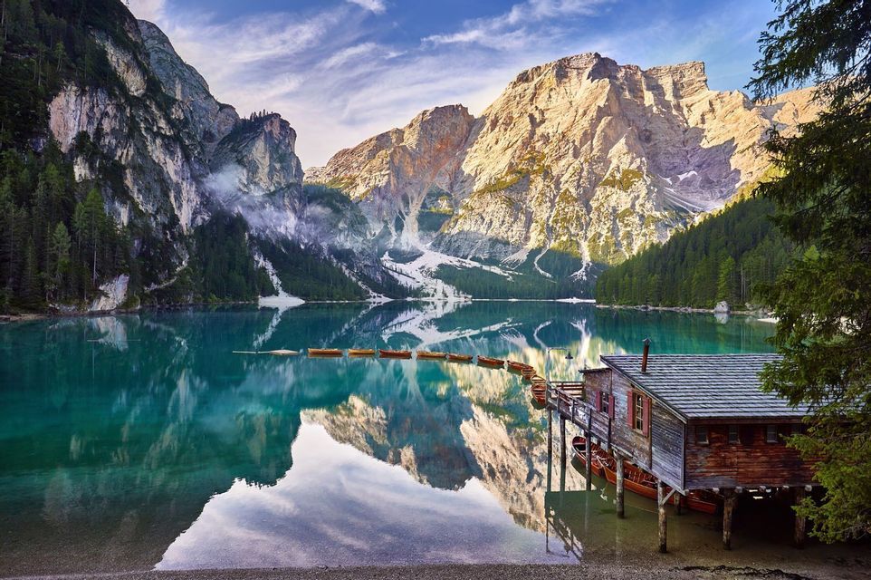 Une cabane à bateaux en bois sur pilotis et une rangée de barques sur un lac turquoise paisible, avec de grandes montagnes et un ciel bleu qui se reflètent dans l'eau.
