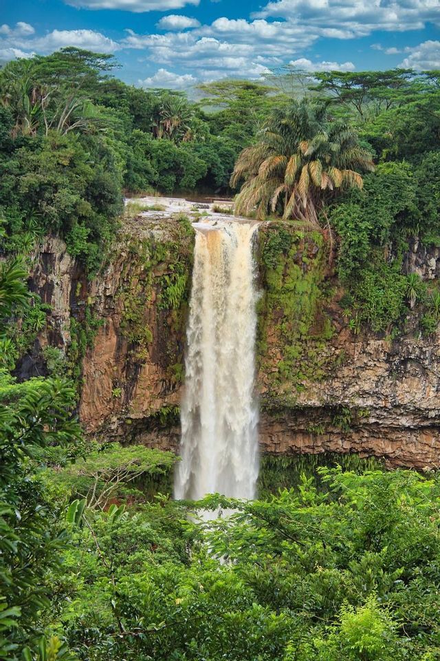 Una potente cascata scende da una scogliera rocciosa muschiosa, circondata da una fitta foresta tropicale sotto un cielo parzialmente nuvoloso.