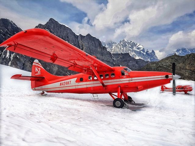 A red propeller plane fitted with skis is parked on a snowy glacier, with a rugged mountain range in the background.