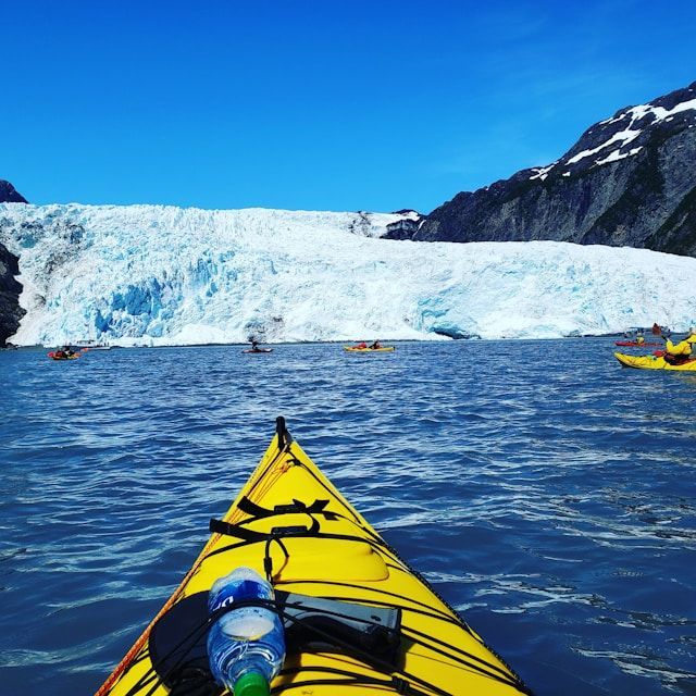First-person view from a yellow kayak as a WeRoad group trip paddles on blue water towards a massive glacier wall under a clear sky.