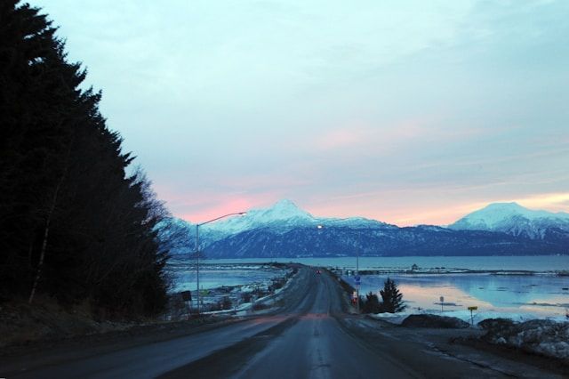 A wet road runs alongside a calm body of water, leading towards snow-capped mountains under a pastel twilight sky.