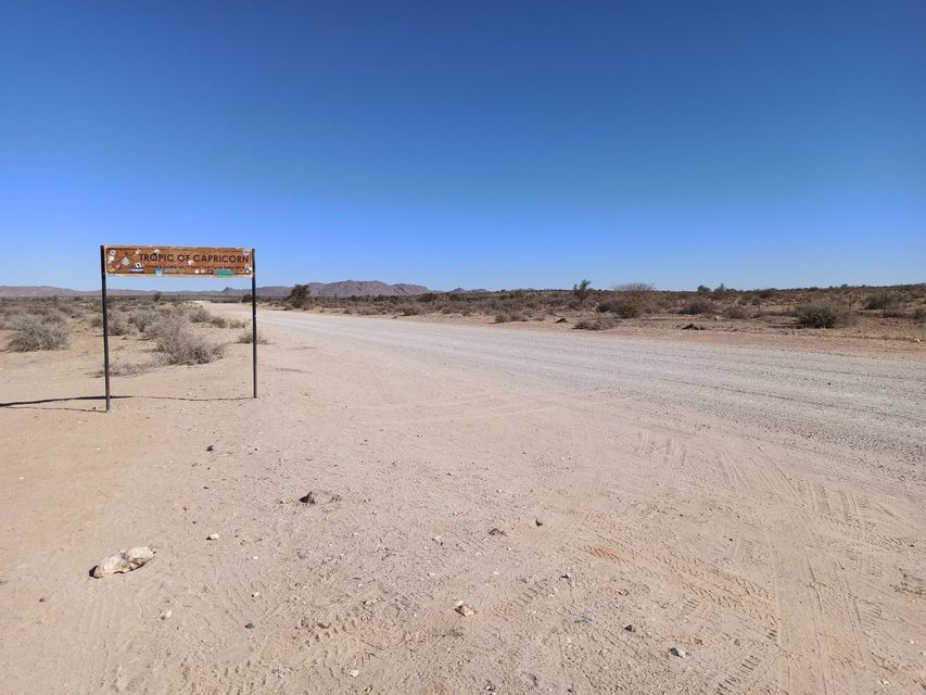 Un cartello 'Tropico del Capricorno' si erge accanto a una lunga strada sterrata in un vasto paesaggio desertico sotto un cielo azzurro.