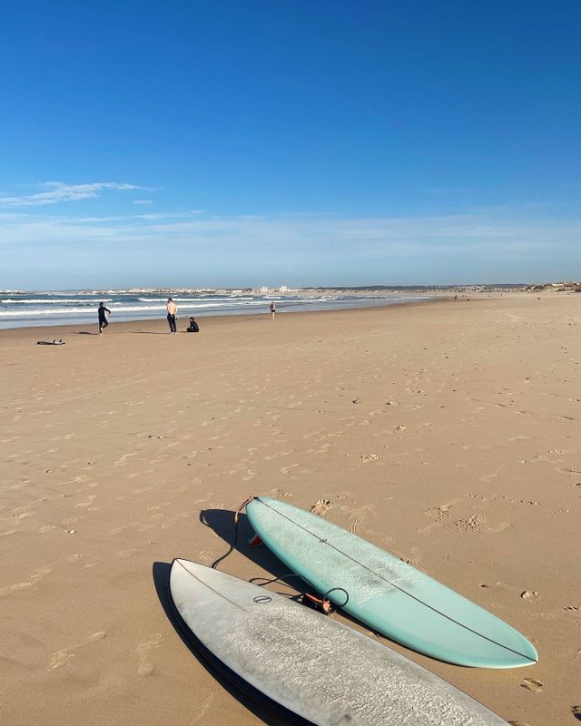 Due tavole da surf su una spiaggia sabbiosa, con persone e l'oceano sullo sfondo, sotto un cielo azzurro e limpido.