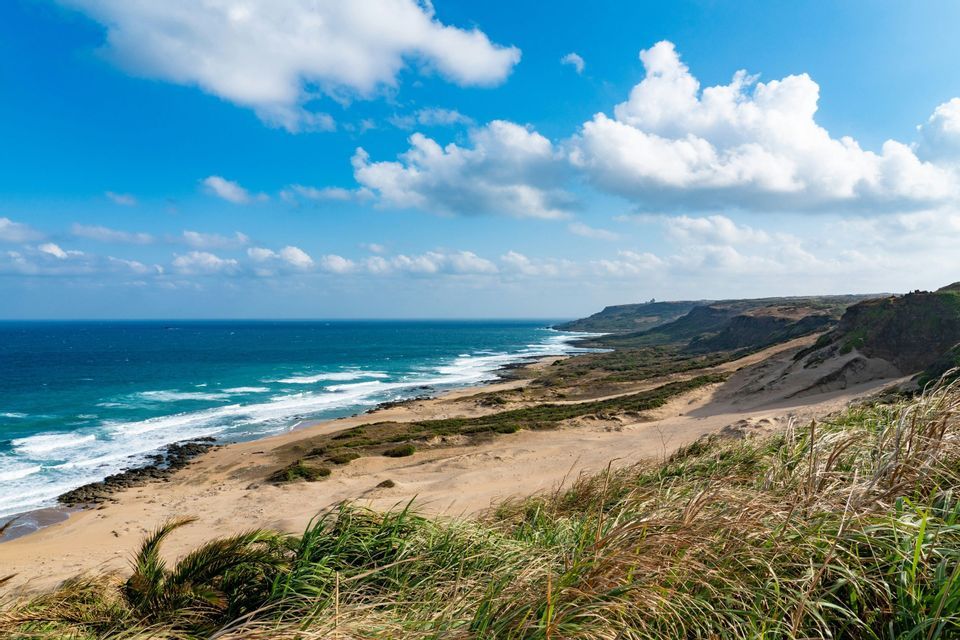 Eine sandige Küstenlinie schlängelt sich entlang eines blauen Ozeans mit sanften Wellen, von einem grasbewachsenen Hügel unter einem hellen, teils bewölkten Himmel aus gesehen.