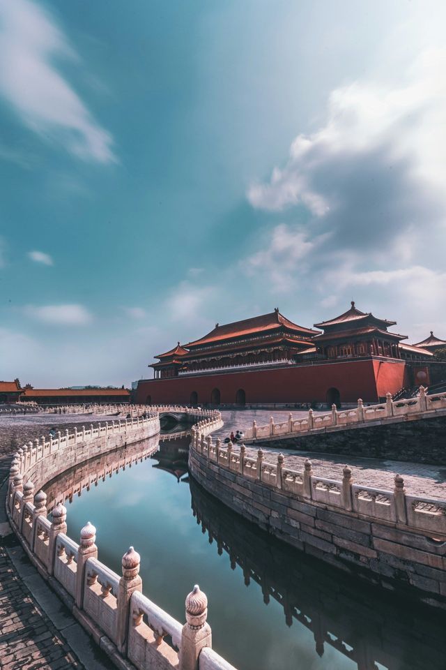 Ein gewundener Wassergraben mit steinerner Balustrade spiegelt einen traditionellen roten chinesischen Palast unter einem gestreiften blauen Himmel wider.