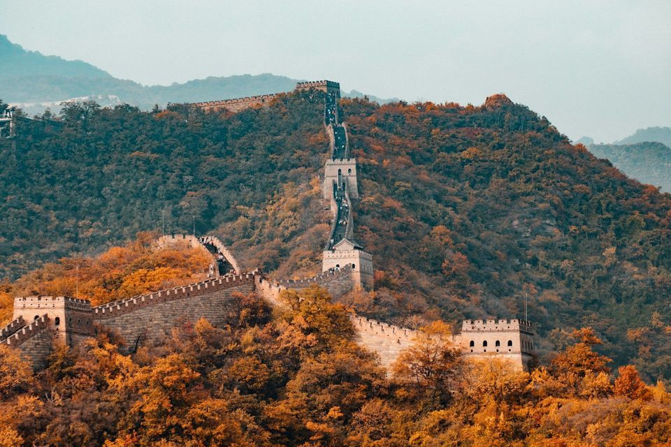 Eine lange Steinmauer mit Wachtürmen zieht sich einen steilen Berg hinauf und hinunter, der von dichten, herbstlich gefärbten Bäumen bedeckt ist.