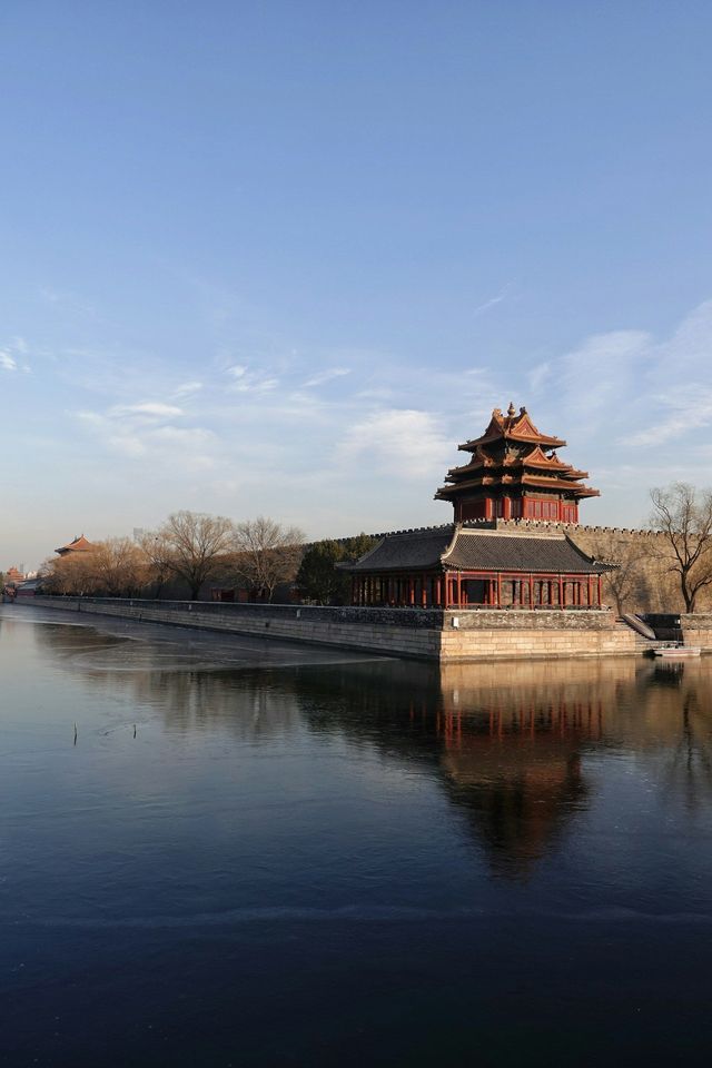 Ein chinesischer Eckturm auf einer Steinmauer spiegelt sich im stillen Wasser eines Wassergrabens unter klarem Himmel.