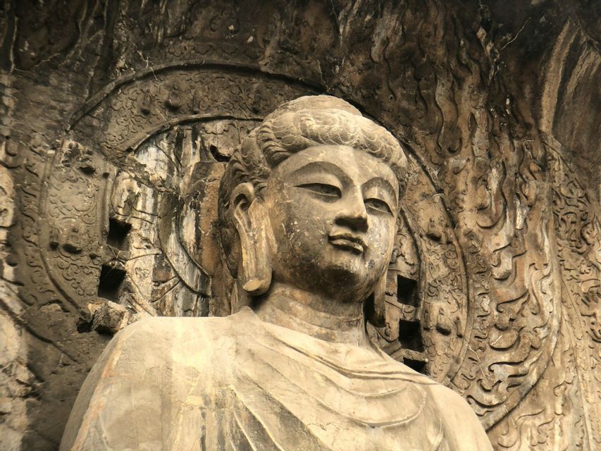 Eine große, verwitterte Steinbuddha-Statue, die in eine Felswand gehauen ist und von aufwendigen Reliefschnitzereien umgeben ist.