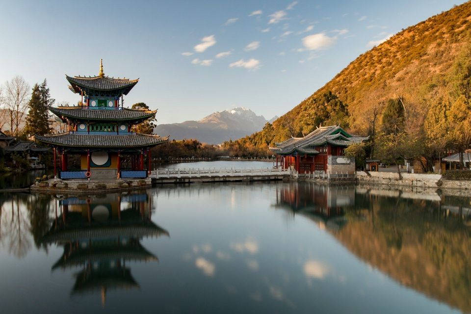Eine traditionelle mehrstufige Pagode und ein Tempel auf einem ruhigen See, mit ihren Spiegelungen im Wasser und einem schneebedeckten Berg im Hintergrund.
