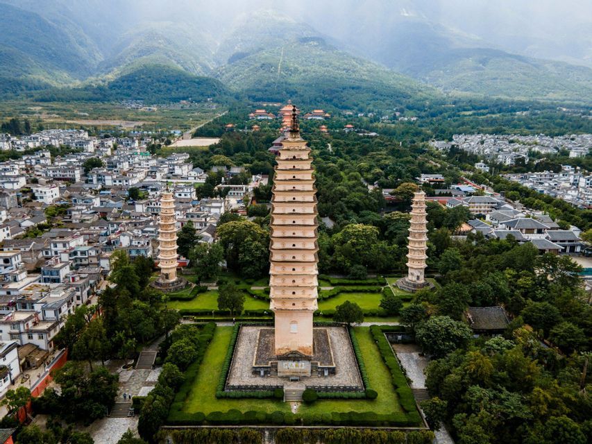 Una veduta aerea di tre pagode tradizionali immerse in un parco verde, circondate da una città ai piedi di montagne nebbiose.
