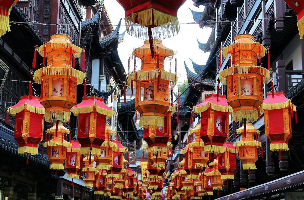 Reihen von roten und orangen chinesischen Laternen mit gelben Quasten hängen über einer Straße zwischen traditionellen Gebäuden.