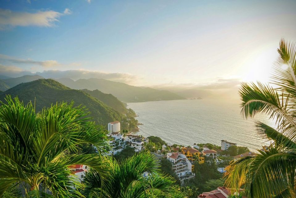An elevated view of a coastal town on a green hillside, framed by palm trees, overlooking the ocean and mountains at sunrise.