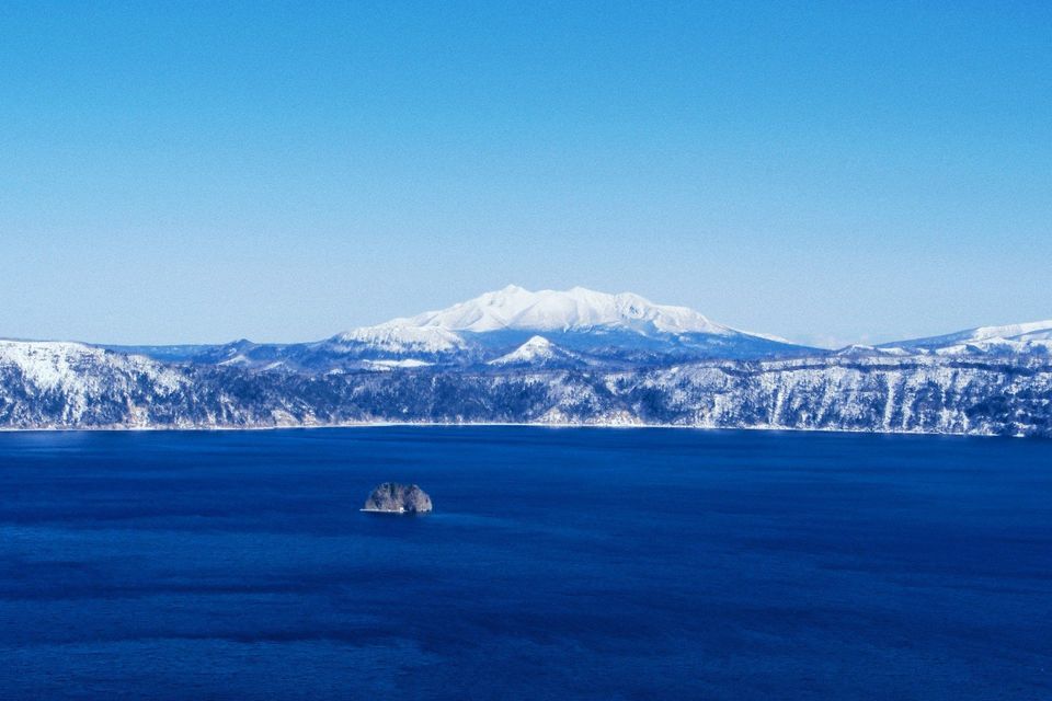 Un'ampia vista di un lago blu profondo con un piccolo isolotto roccioso, sullo sfondo montagne innevate sotto un cielo limpido.