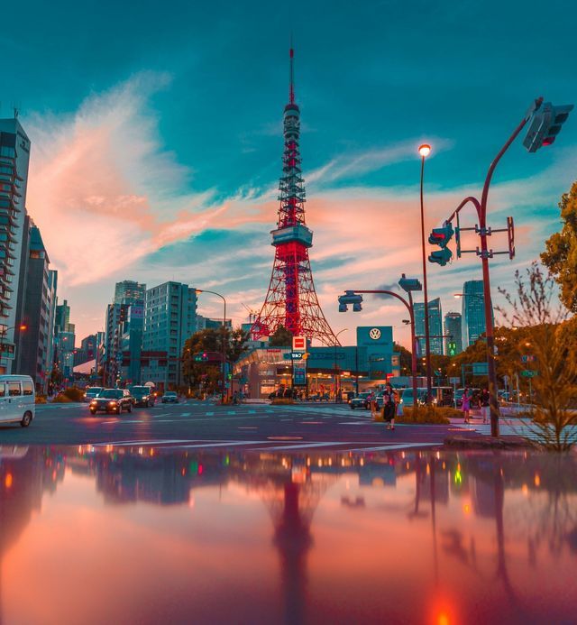 La Torre di Tokyo rossa si erge su un paesaggio urbano al tramonto, il suo riflesso visibile su una strada bagnata in primo piano.