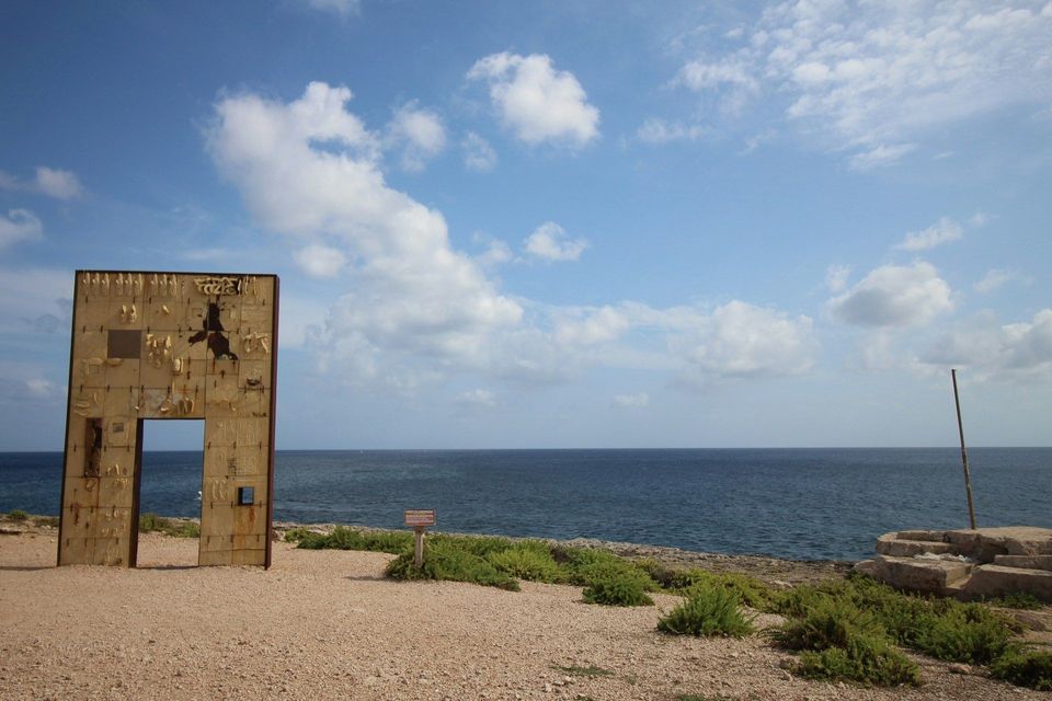 Una grande installazione artistica, erosa dalle intemperie e a forma di portale, si erge su una costa ghiaiosa che si affaccia sul mare, sotto un cielo azzurro con nuvole bianche.