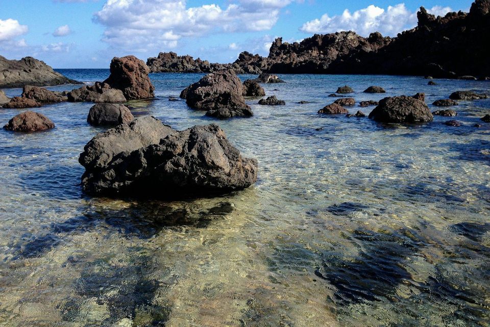 Rocce vulcaniche scure emergono da acque limpide e poco profonde lungo una costa sotto un cielo parzialmente nuvoloso.