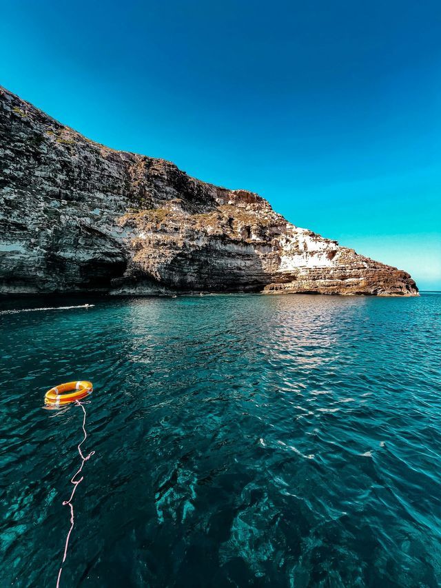 Un salvagente arancione galleggia sul mare turchese intenso accanto a una scogliera rocciosa sotto un cielo azzurro limpido.