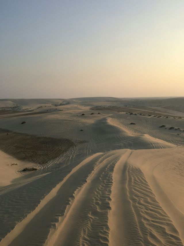 Tracce di pneumatici si snodano tra le vaste dune di sabbia del deserto sotto un cielo velato al tramonto.