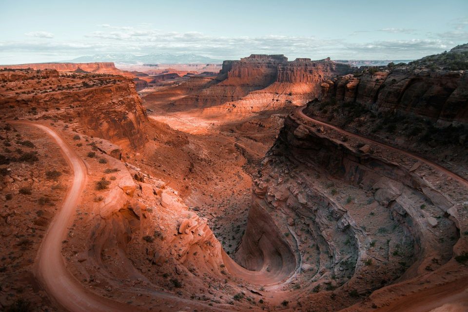 Una strada sterrata tortuosa attraversa un vasto canyon di roccia rossa sotto un cielo azzurro pallido.