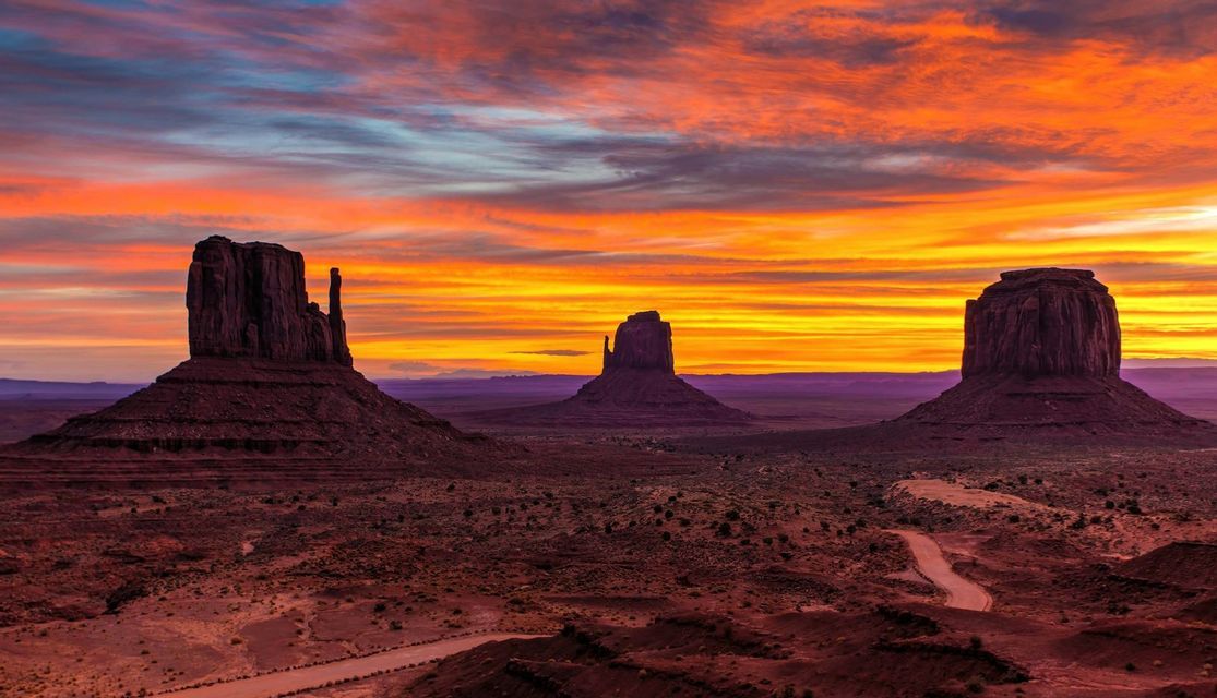 Tre grandi butte di arenaria si ergono in una valle desertica sotto un cielo striato di arancione, giallo e rosso dal tramonto.