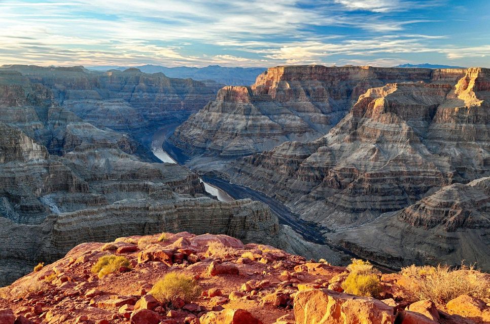 Un vasto canyon stratificato con un fiume sottostante, ammirato da un promontorio roccioso rosso all'alba, sotto un cielo parzialmente nuvoloso.