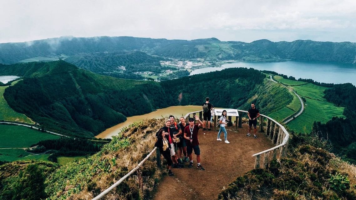 Un groupe WeRoad en voyage pose pour une photo sur un point de vue panoramique surplombant une vallée avec deux lacs et des collines verdoyantes.