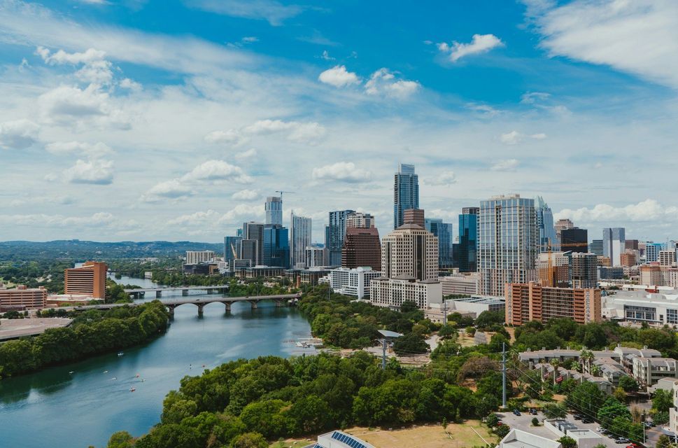 Uno skyline urbano con grattacieli si staglia dietro un ampio fiume, circondato da una vegetazione lussureggiante, sotto un cielo parzialmente nuvoloso.