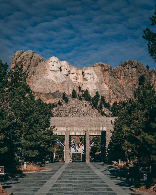 Il Monte Rushmore National Memorial visto dal vialetto d'ingresso in pietra, fiancheggiato da pini sotto un cielo azzurro.