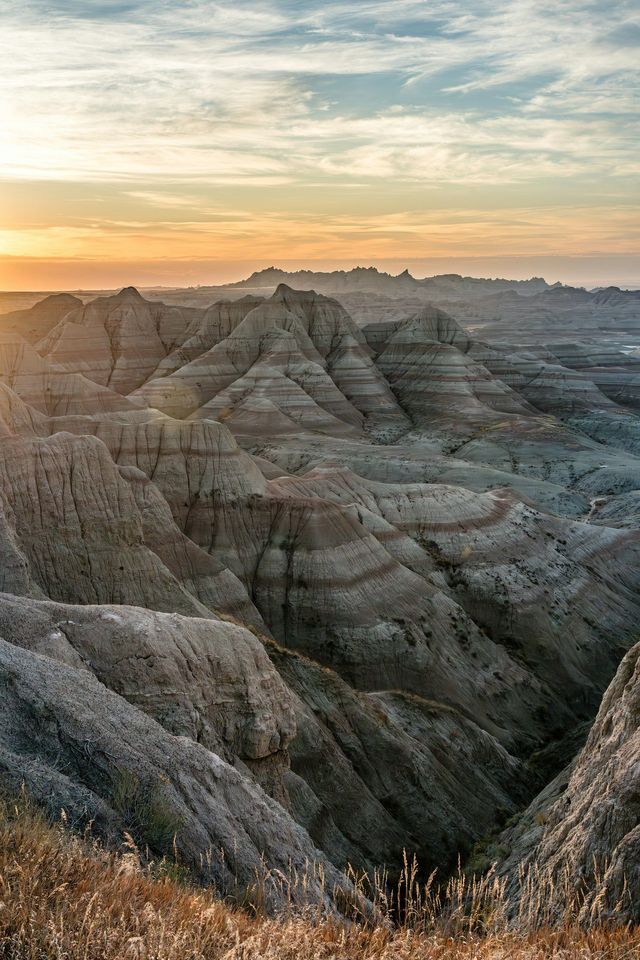 Un vasto paesaggio di formazioni rocciose stratificate (calanchi) illuminato dalla calda luce di un tramonto sotto un cielo parzialmente nuvoloso.