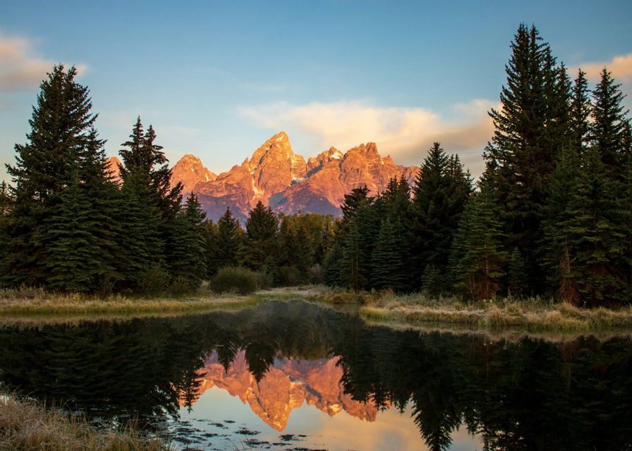 Cime montuose frastagliate illuminate dal sole si riflettono in un lago immobile, con una fitta foresta sempreverde in primo piano.