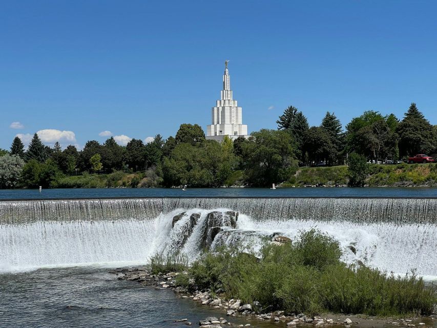 Un'ampia cascata scende lungo un fiume, con un tempio bianco a più livelli che si erge sopra gli alberi sulla sponda opposta sotto un cielo azzurro.