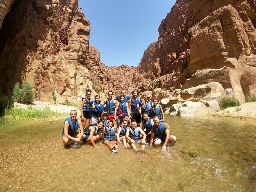 A WeRoad group trip wearing life vests poses for a photo in a shallow river at the bottom of a sunny canyon.