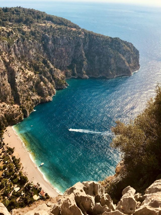 Vista dall'alto di un motoscafo che solca l'acqua turchese di una cala, con una spiaggia sabbiosa adagiata ai piedi di una grande scogliera.