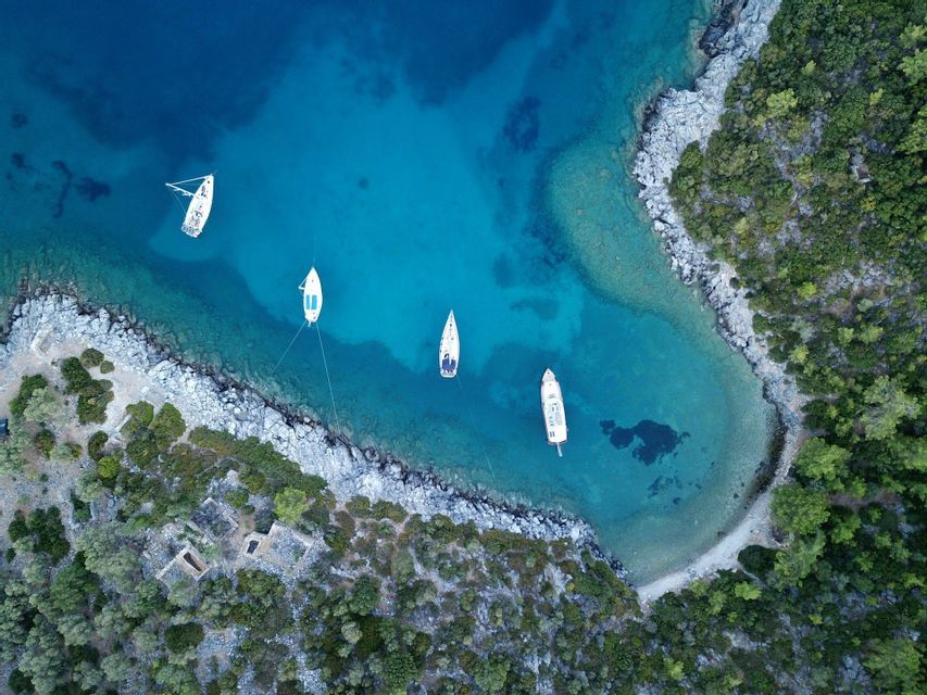 Vista aerea di barche a vela ancorate in una caletta cristallina e turchese, adiacente a una costa rocciosa e boscosa.