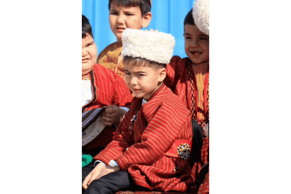 Un bambino, con una giacca tradizionale a righe rosse e un cappello di lana bianco, siede in un gruppo di bambini, guardando la fotocamera.