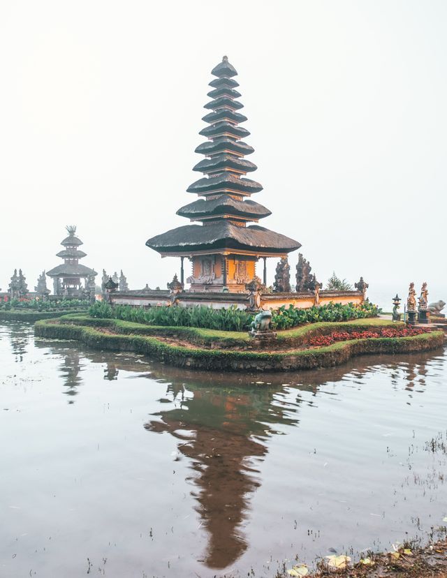 Un tempio asiatico a più livelli su una piccola isola circondata da acque calme, con il suo riflesso visibile in una giornata nebbiosa.