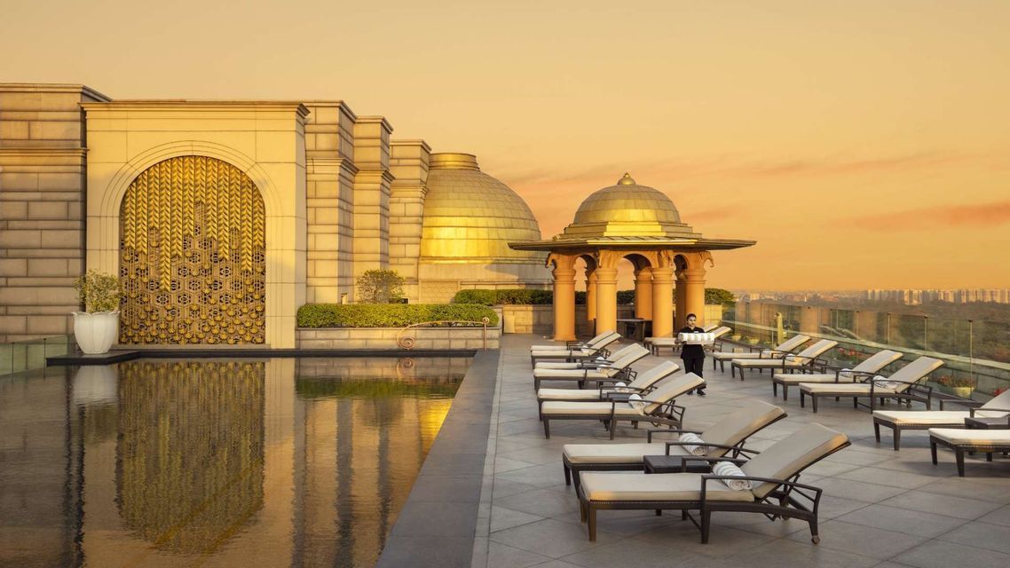 Un cameriere porta un vassoio accanto a una piscina panoramica con sedie a sdraio, vicino a un edificio sontuoso con cupole dorate sotto un cielo al tramonto.