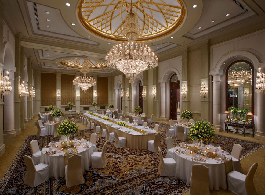 Una sala da ballo sontuosa con grandi lampadari di cristallo sospesi sopra tavoli da pranzo apparecchiati per un evento formale.