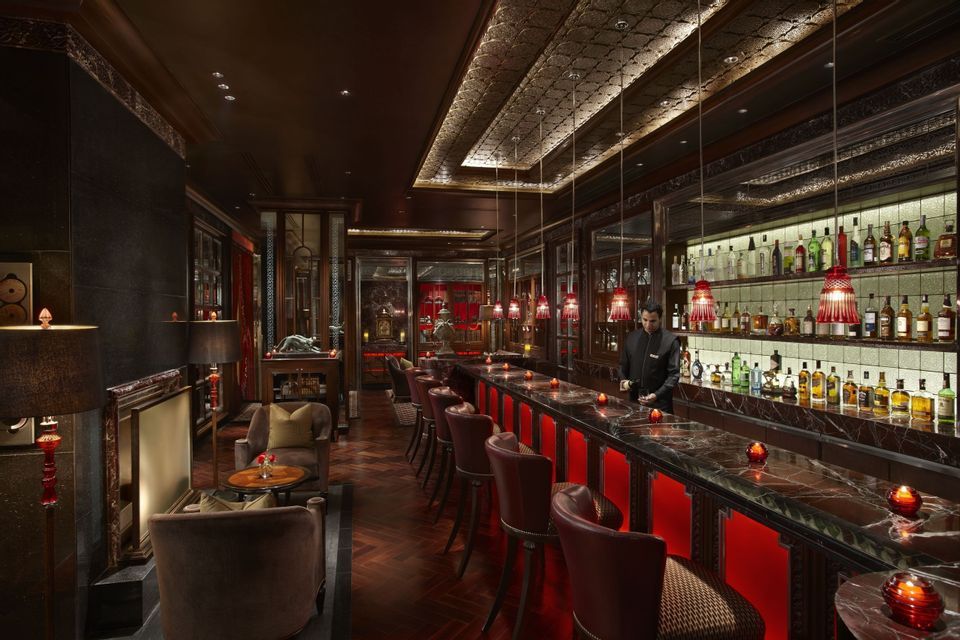 Ein Barkeeper steht hinter einer langen Marmortheke in einer gedämpft beleuchteten, gehobenen Bar mit roten Hockern und aufwendiger Dekoration.