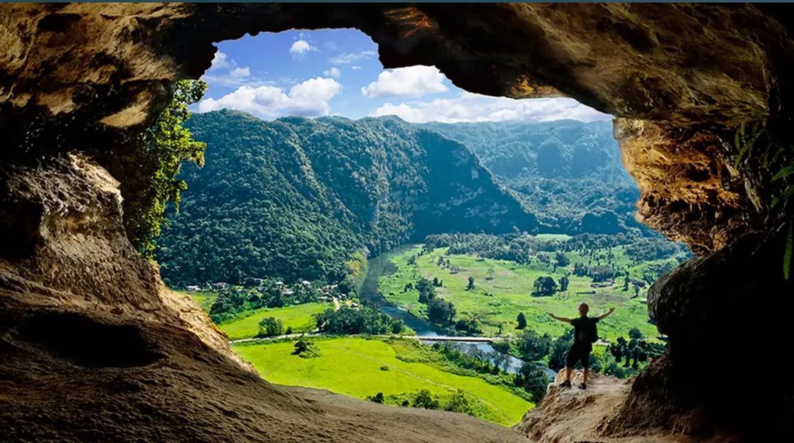 A person with arms outstretched stands at the mouth of a cave, looking out over a lush green valley with mountains and a river.