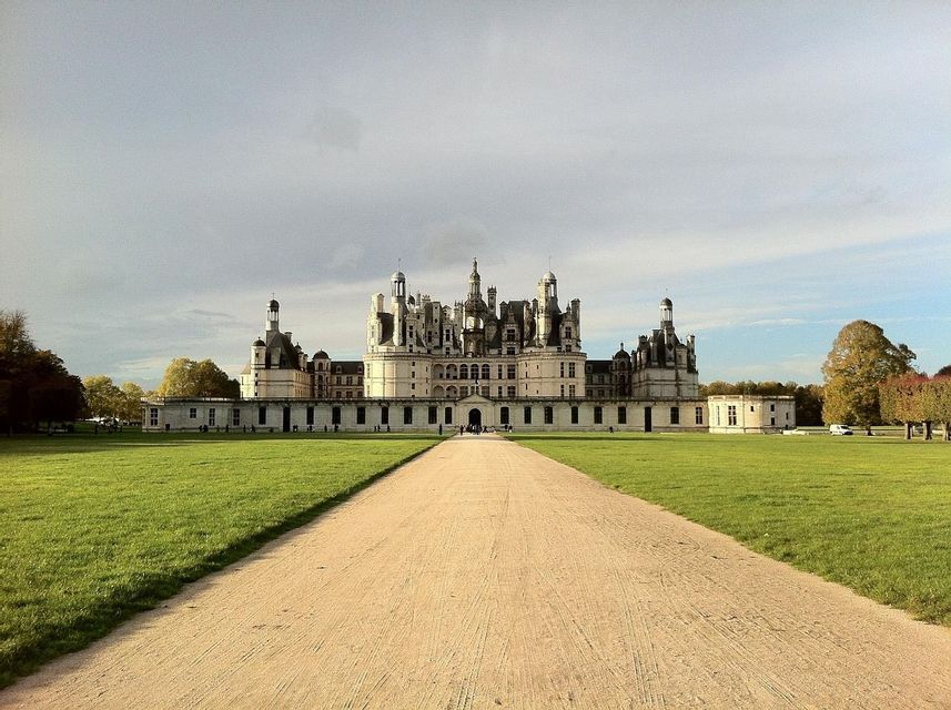 A long, unpaved path leads across a vast green lawn to a large, ornate white castle with many towers under a partly cloudy sky.