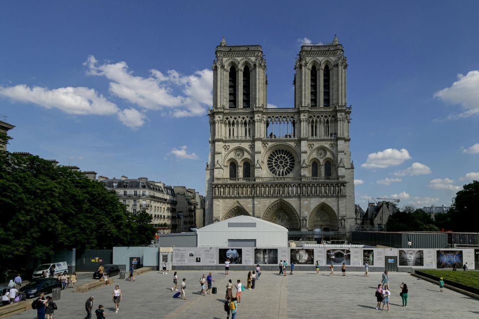 The facade of Notre Dame Cathedral on a sunny day, with people walking in the large plaza in front of the reconstruction site.