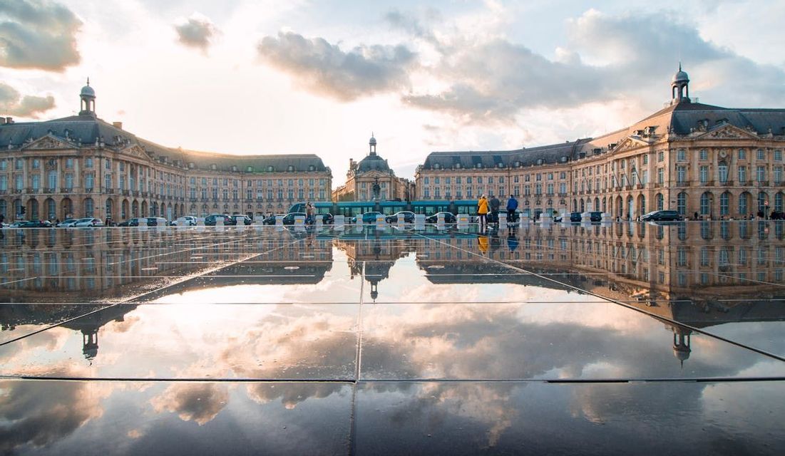 A grand historical building and cloudy sky are reflected in the wet surface of a large city plaza at sunset.