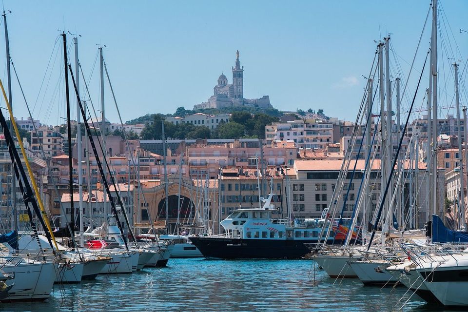 A bustling harbor filled with sailboats and a tour boat, with a city and a prominent basilica on a hill in the background.