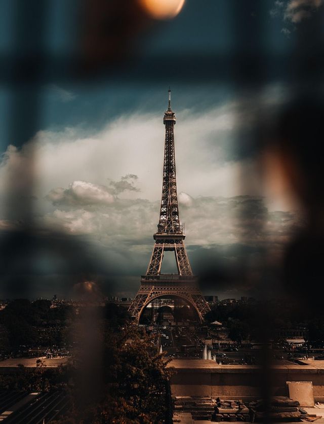 A view of the Eiffel Tower seen through a blurred foreground, set against a dark, cloudy sky over the cityscape.