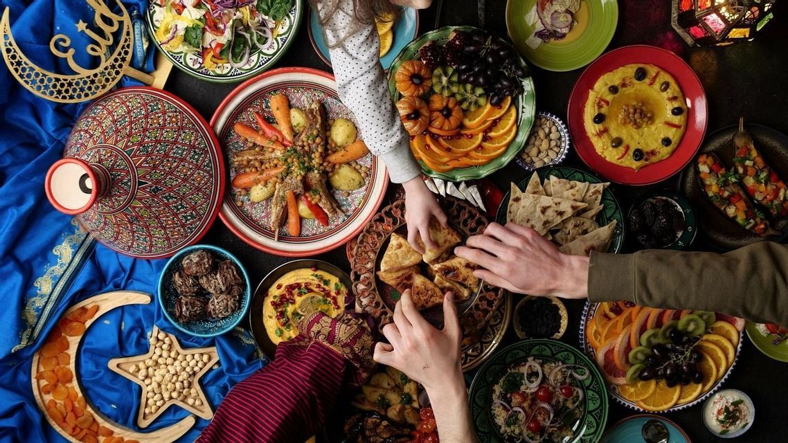 Vista cenital de una mesa festiva con tagine, hummus, frutas y pan de pita, mientras varias manos se acercan a la comida.