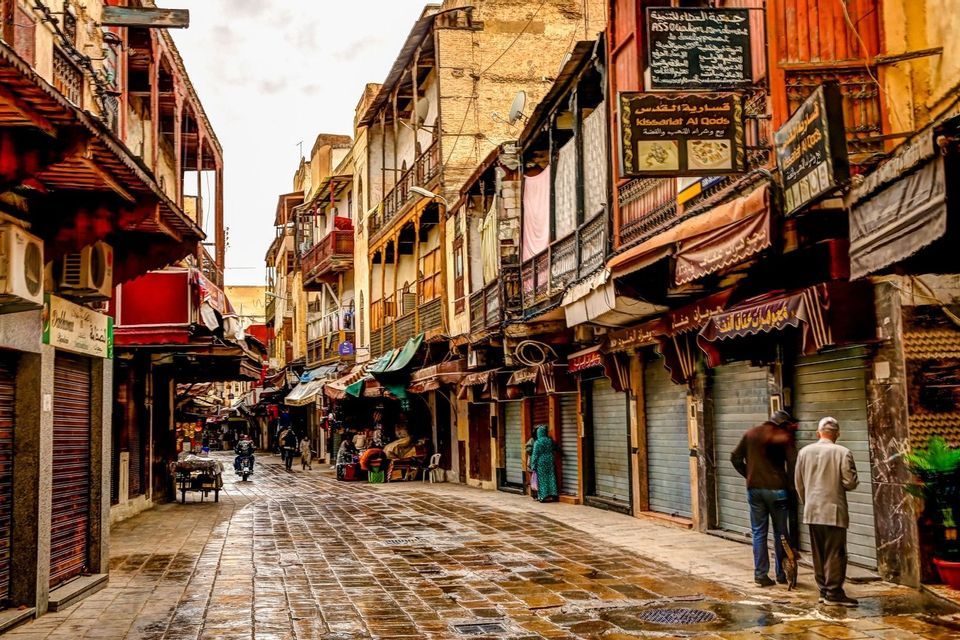 Una calle adoquinada húmeda en un mercado estrecho, con gente pasando junto a edificios de varios pisos y tiendas cerradas.