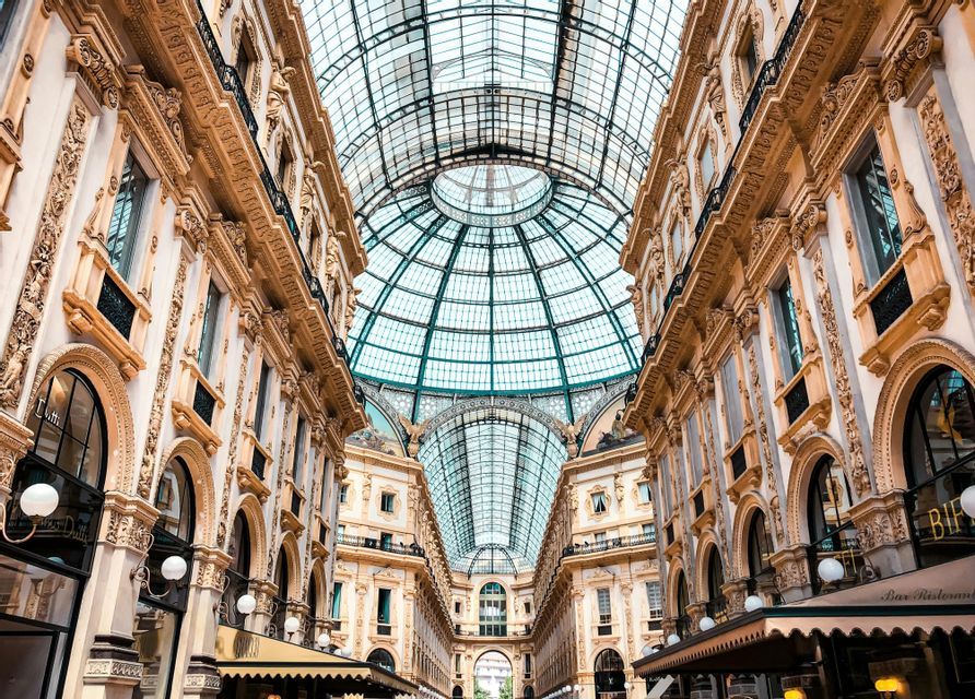 A low-angle view looking up at the arched glass and steel roof of an ornate, multi-story shopping arcade with classical architecture.