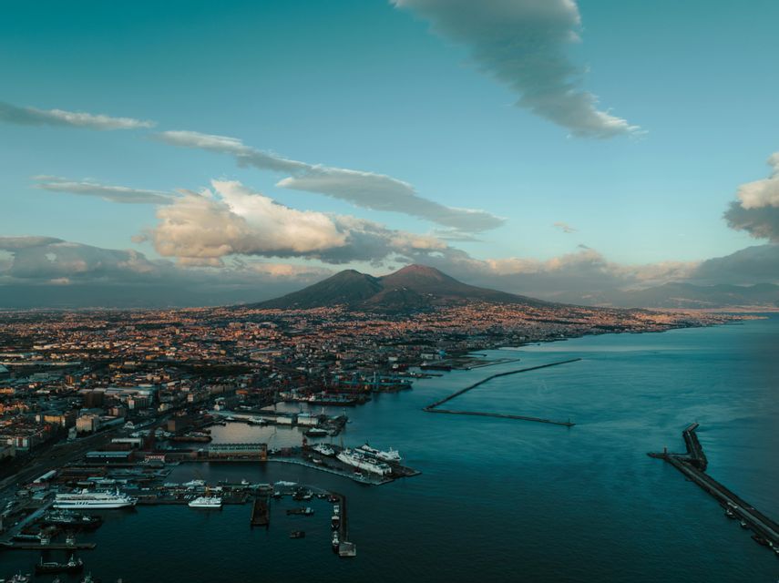 An aerial view of a sprawling coastal city and its harbor, with a large, two-peaked mountain in the background under a partly cloudy sky.